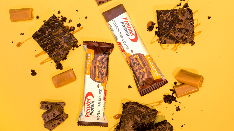 Two Premier Protein chocolate protein bars on a yellow background with chocolate pieces scattered around.