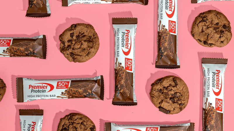 Premier Protein bars and cookies on a pink background