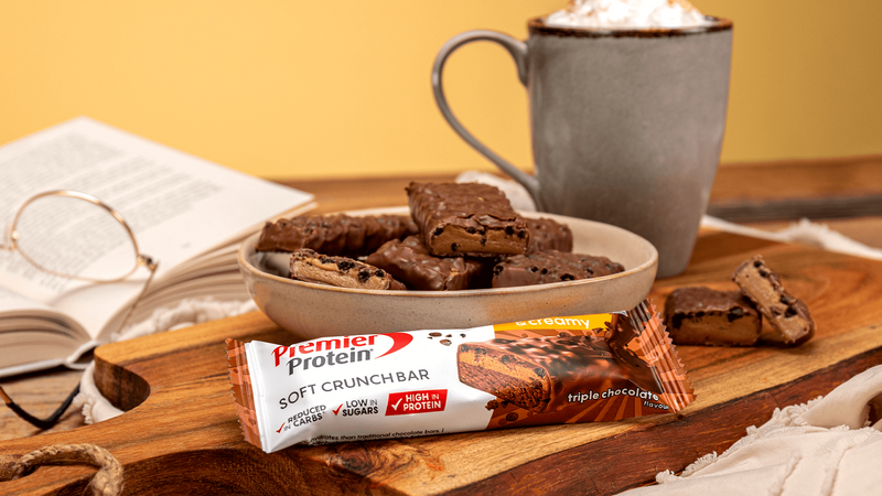 Premier Protein soft crunch bar on a wooden tray with brownies and a mug of hot chocolate.