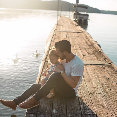 Man and child sitting on a wooden dock by a lake with ducks in the water.