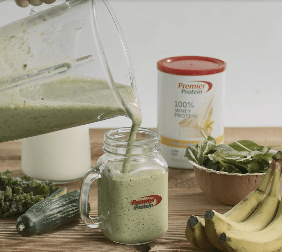 Green smoothie being poured into a jar with Premier Protein powder and ingredients like bananas and spinach in the background.