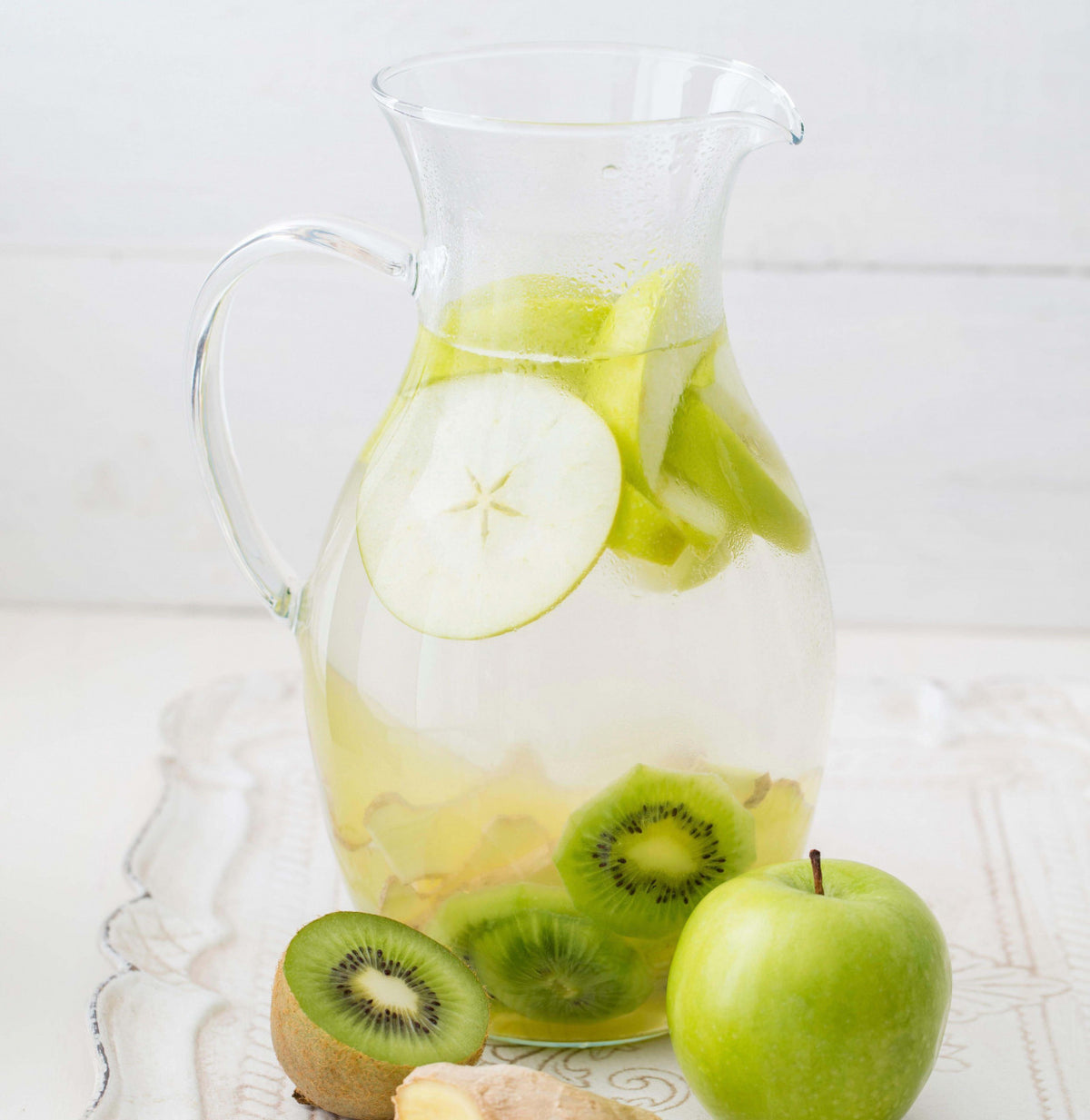 Clear pitcher filled with green apple slices and kiwi pieces on a white surface