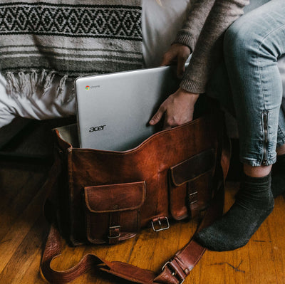 Person holding a laptop in a brown leather bag on a wooden floor.