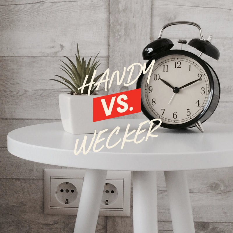 Alarm clock and plant on a table with 'HANDY VS. WECKER' text.