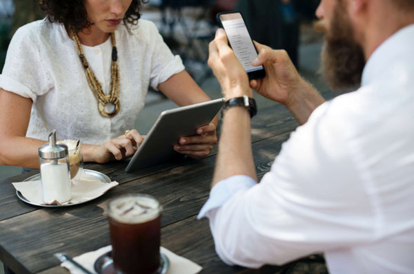 Two people sitting at a table with drinks and digital devices.